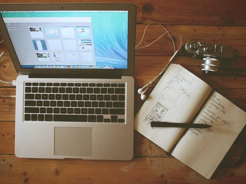 Laptop with open notebook of handwritten sketches and notes, a pen, camera and earbuds arranged on a wooden tabletop.