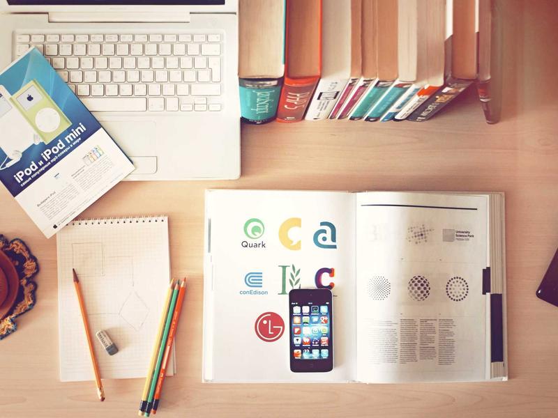 Work desk with laptop, pencils, books, open book showing logos 'Quark', 'conEdison', 'LG' and pamphlet reading 'iPod iPod mini'.