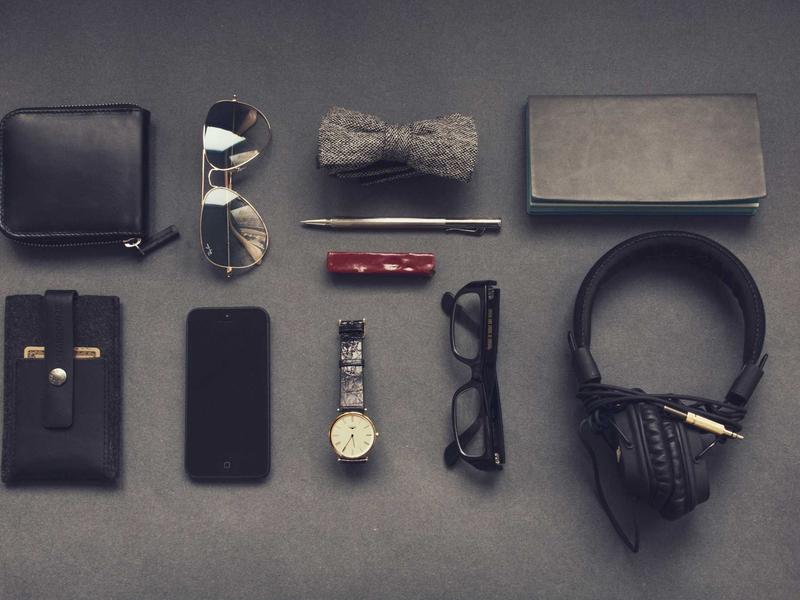 Neatly arranged flat-lay of men's everyday accessories: wallet, sunglasses, bow tie, pen, watch, phone, glasses, headphones, and notebook.
