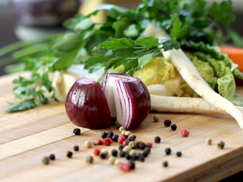 Halved red onion and scattered multicolored peppercorns on a wooden cutting board with parsley and root vegetables behind.