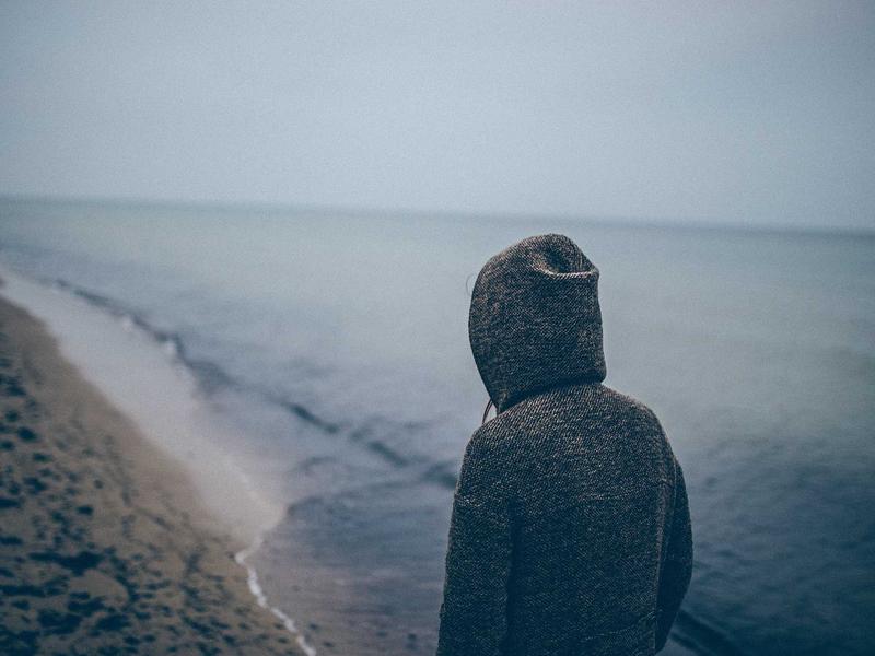Solitary hooded figure facing the calm sea on a misty, empty beach, hinting at quiet reflection.