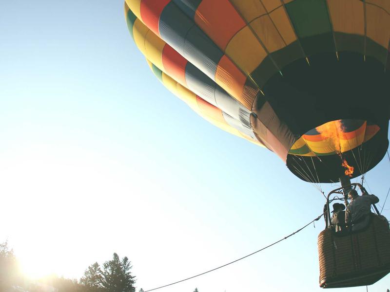 Colorful hot air balloon ascending at sunrise with two people operating the burner above a treeline.
