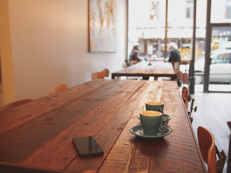Rustic bakery table with two espresso cups and a phone in the foreground, patrons talking near the front window.