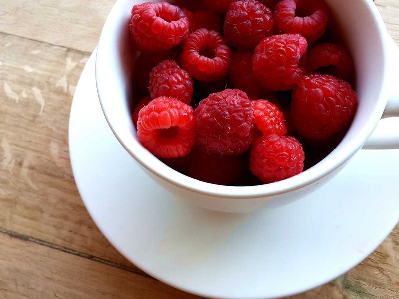 White teacup on a saucer filled with bright red raspberries resting on a wooden surface.
